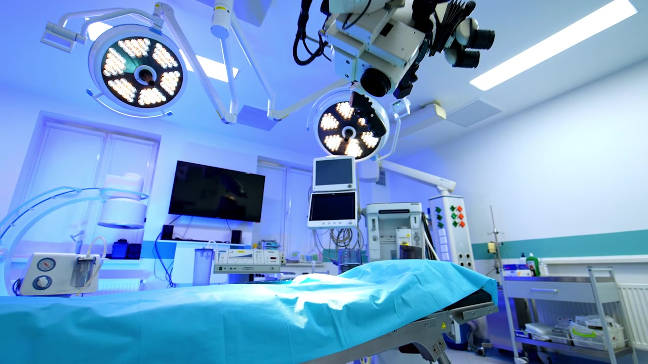 Entering the empty doors to a large modern surgical room. Low angle view at the equipment. Approaching the empty operating table lit with light from lamps on the ceiling.