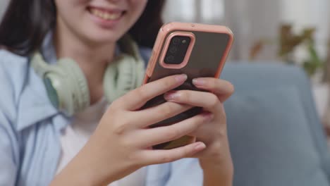 cerca de las manos de una adolescente asiática jugando con su teléfono inteligente mientras está sentada en el sofá en la sala de estar en casa