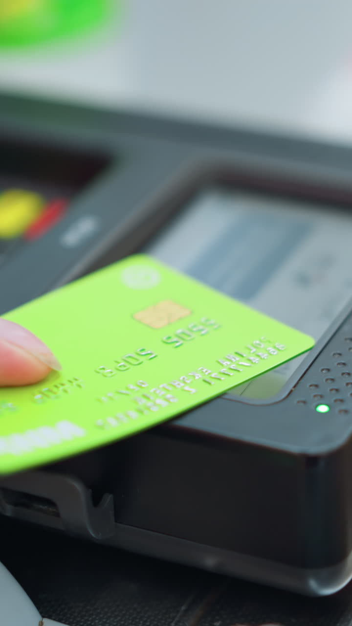vista a mano de alguien con un vestido rosa colocando una tarjeta verde en un lector de tarjetas para hacer un pago en un centro comercial bien iluminado, con otros equipos parcialmente visibles en el fondo, ligeramente borrosos