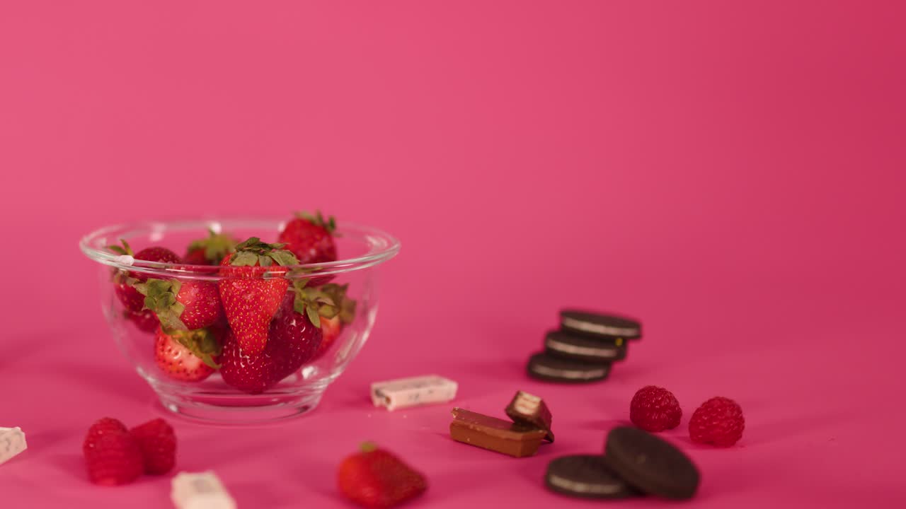Hand removes strawberries, chocolate, and candy from glass bowl on pink background, soft lighting
