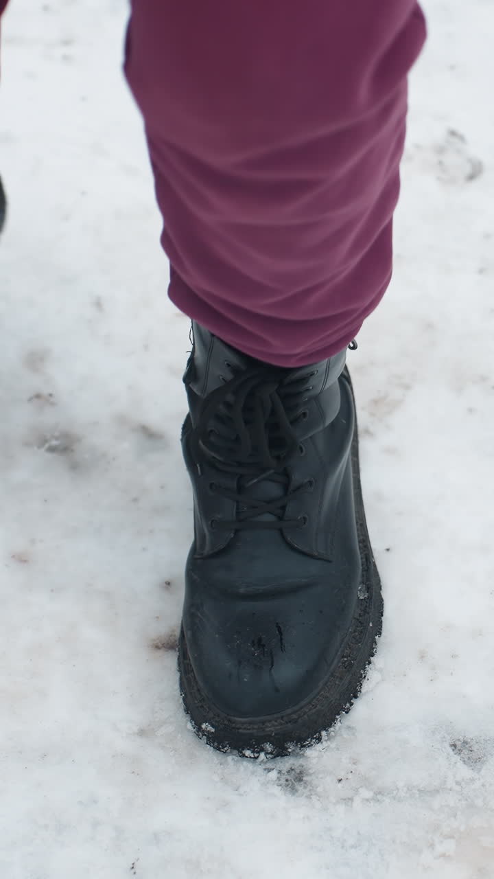 vista de ángulo inferior de un individuo con botas negras saltando juguetonamente en el suelo cubierto de nieve con follaje seco esparcido, centrarse en el calzado, el movimiento y el entorno de invierno