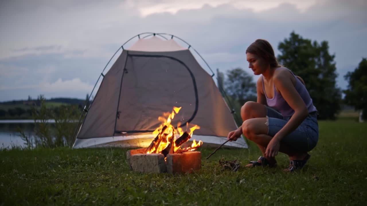 Kvinde der sidder på hug i græsset og lægger pinde på et brændende lejrbål nær telt og rolig sø i skumringen, der holder flammen ved lige og fanger den fredfyldte udendørs campingatmosfære i Savci, Slovenien.
