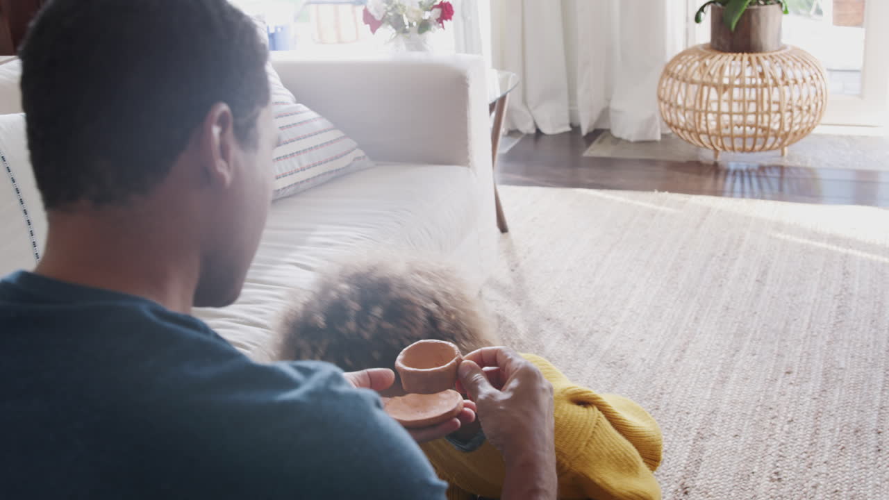 Pre-teen African American girl giving her dad a cup and saucer she&rsquo;s made from clay, over shoulder view