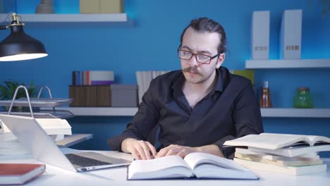 Focused-Economist-reading-book,-looking-for-information-using-laptop.