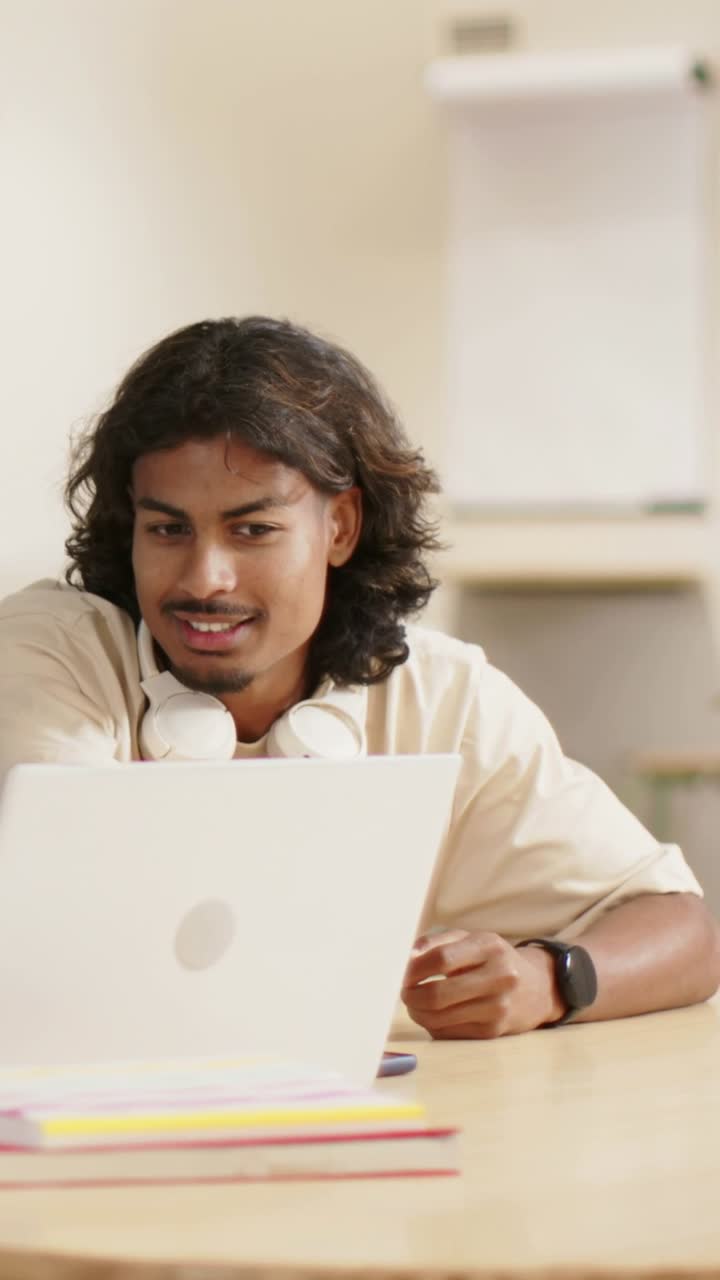 Vertical video: Watching laptop, man smiling and nodding at study desk with headset, in light shirt