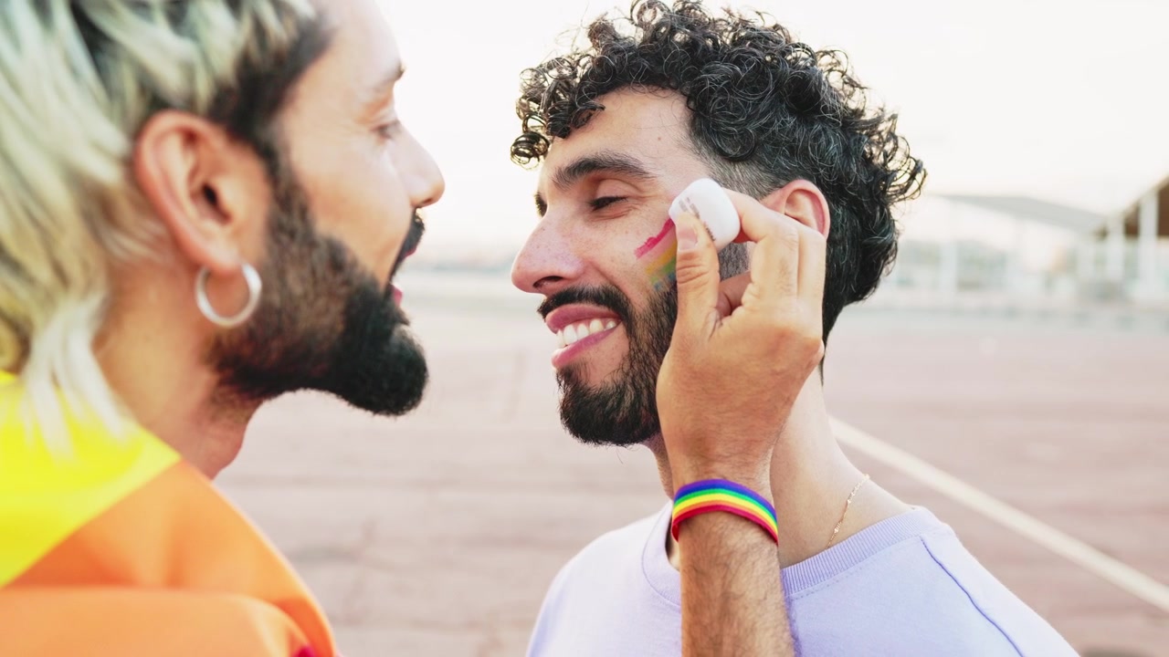 Uomo gay applica trucco arcobaleno sulla guancia del partner.