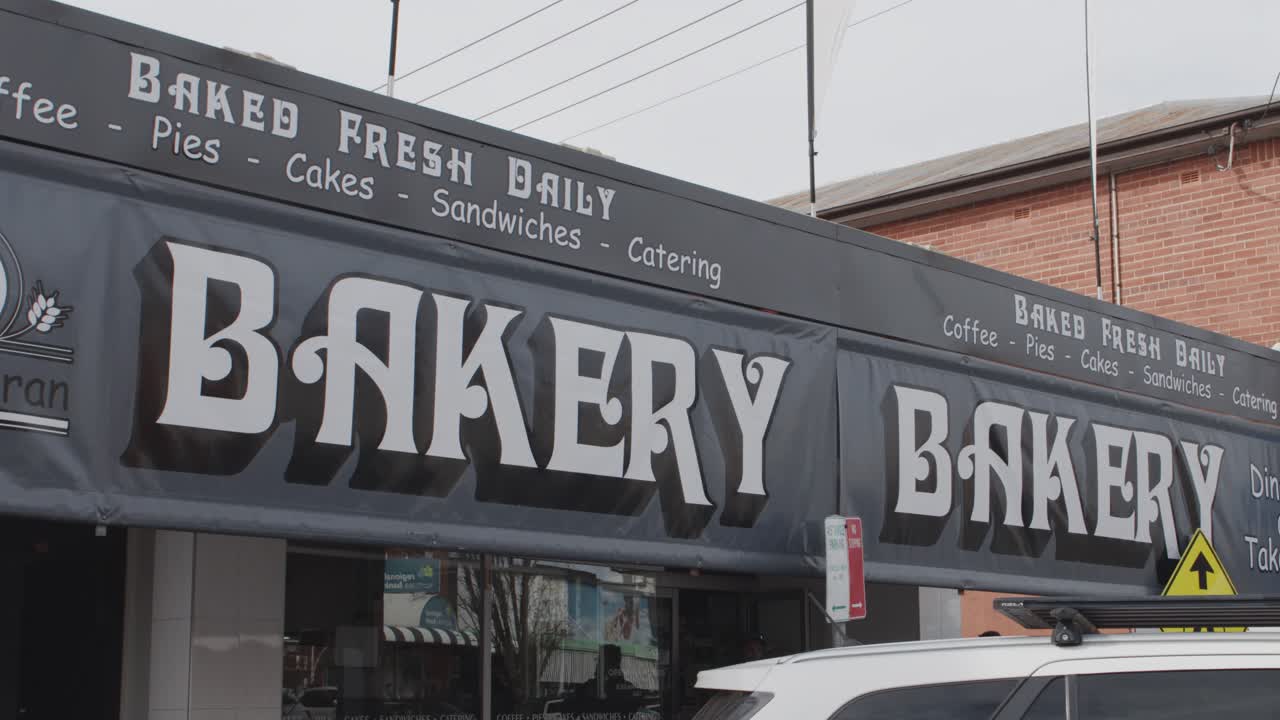 White SUV passes bakery storefront on cloudy day, camera pans right along quiet Australian street