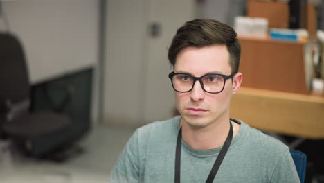 focused young man wearing glasses working in office environment, looking at computer screen, concentrating on task. professional setting with modern workspace, technology to detail