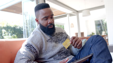 African-american-man-using-tablet-and-credit-card-in-sunny-living-room,-slow-motion