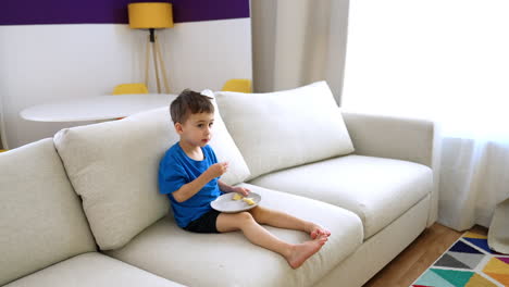 Child enjoying snack on a cozy couch. A young boy in a blue shirt sits comfortably on a soft couch, eating a snack in a brightly lit living room