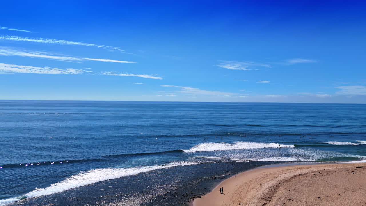 Serene blue waterscape of the Pacific Ocean on sunny day. Some surfers are in the water. People walk by the sandy beach of Malibu, California, USA. Aerial view
