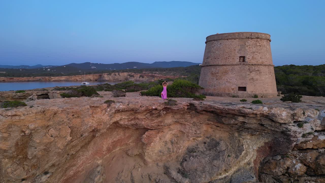 Carefree woman in pink dress in Yoga tree pose enjoying freedom and breathtaking view on Ibiza island near Torre des Savinar at sunset. Smooth aerial view flight speed ramp hyper motion time lapse