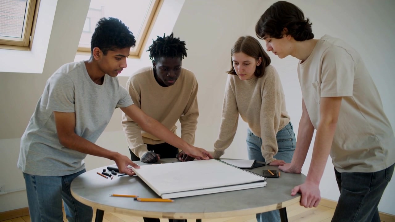 Opening sketchbook, leftmost student pointing at page at loft table, planning design with pencils
