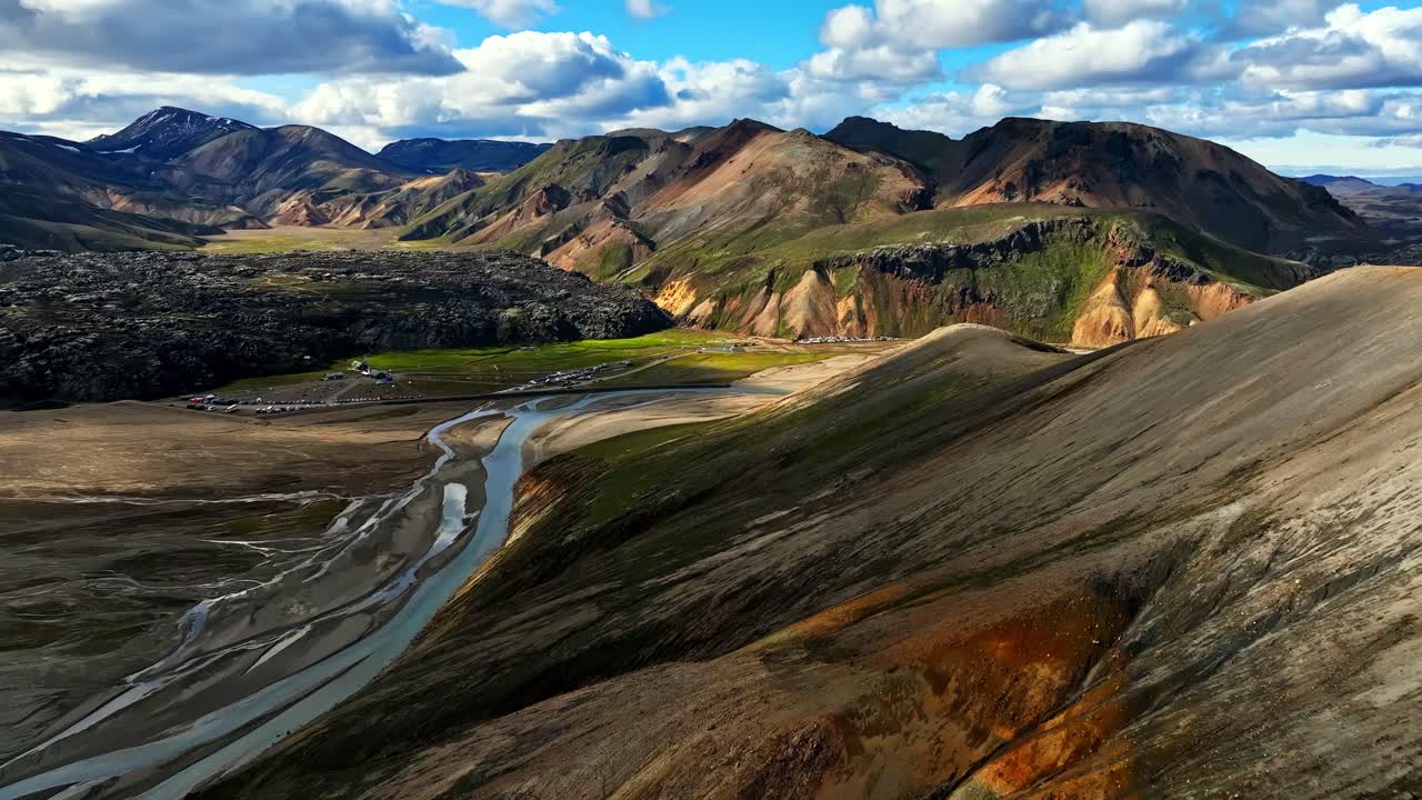 Soft sunlight highlights a ridged Icelandic mountainside as a braided river winds through a wide valley below, revealing striking earthy colors and sweeping highland depth