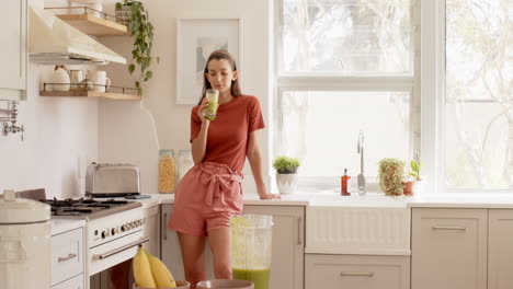 woman in kitchen enjoying morning sunlight, standing near counter with smoothie