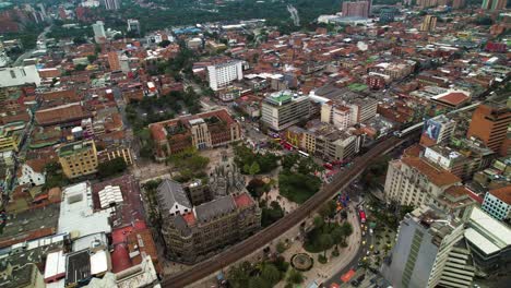 Ein-Zug-Fährt-Durch-Die-Stadt-Medellin-Entlang-Der-Überführung-Um-Die-Christliche-Kirche-Herum,-Mit-Hohen-Wolkenkratzern-Im-Hintergrund