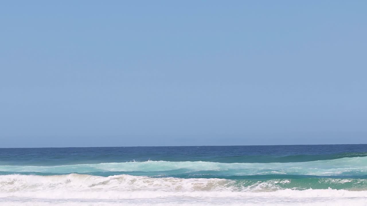 A series of gentle ocean waves roll steadily under a bright, clear blue sky.