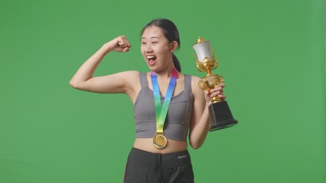 asian woman with a gold medal and trophy flexing her bicep and smiling to camera on green screen background in the studio