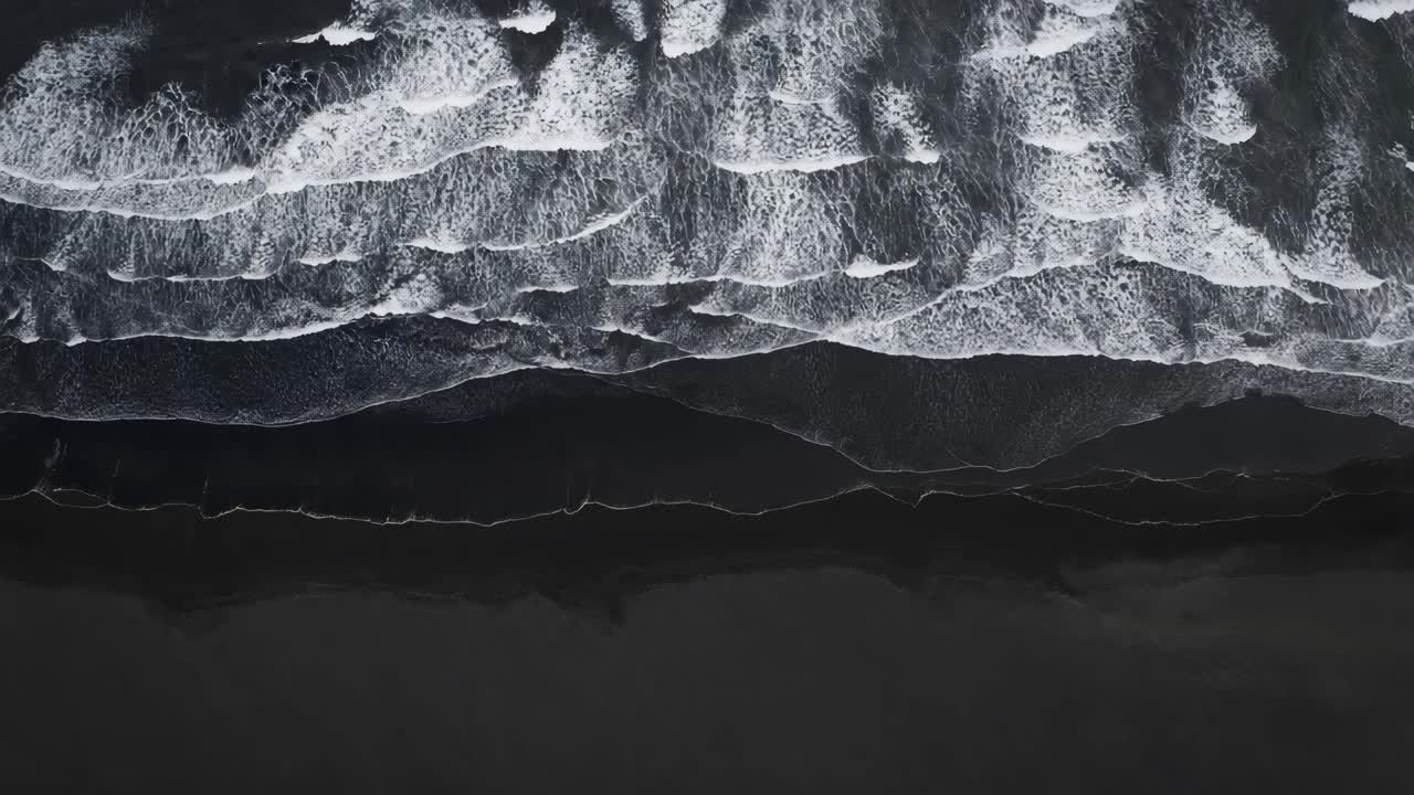 Aerial view of ocean waves crashing on a dark beach, creating a mesmerizing pattern