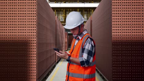side view of asian male engineer with safety helmet using smartphone while standing with red brick packed in stacks are stored