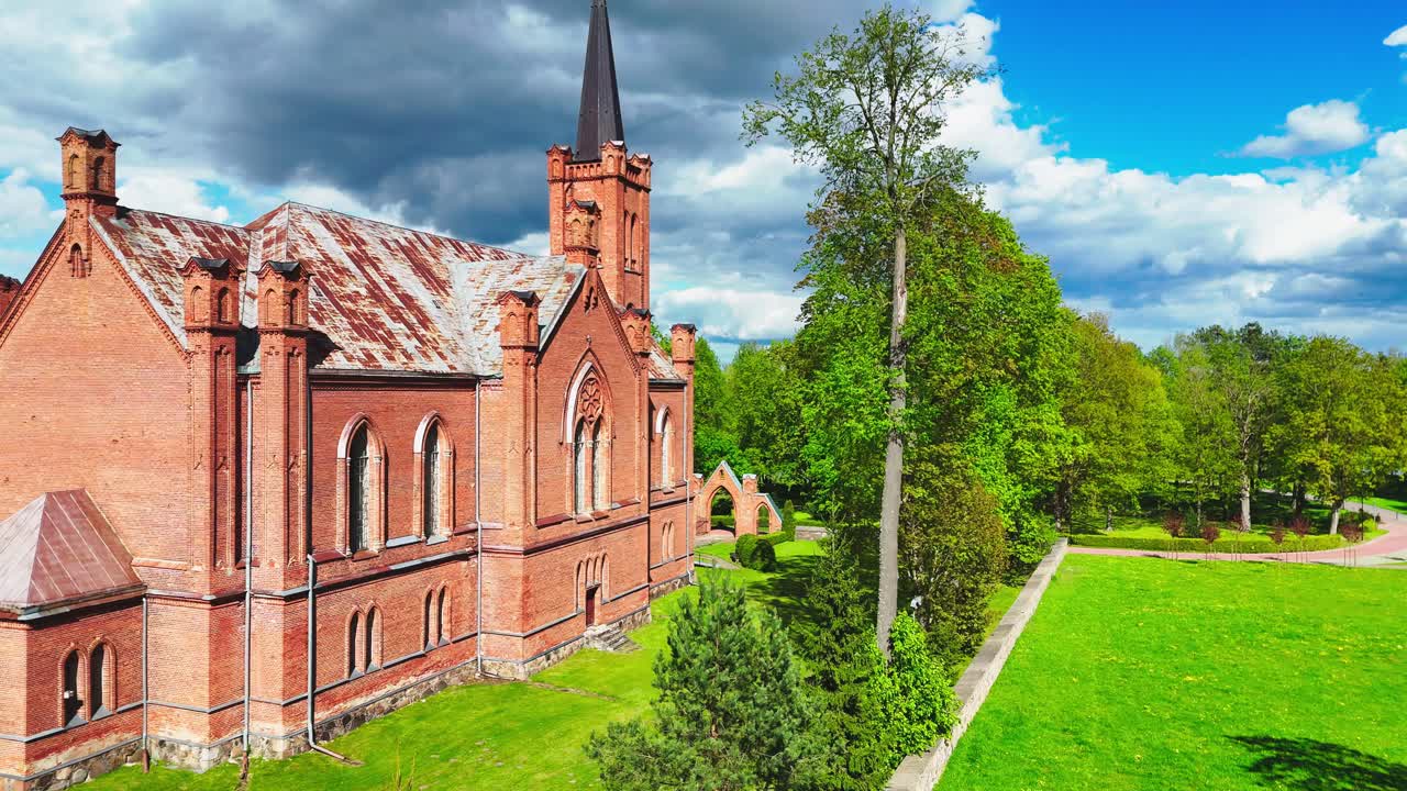 A historic red church with a pointed spire and weathered roof sits beside a tree-lined path in a green spring landscape under dramatic clouds and blue sky.