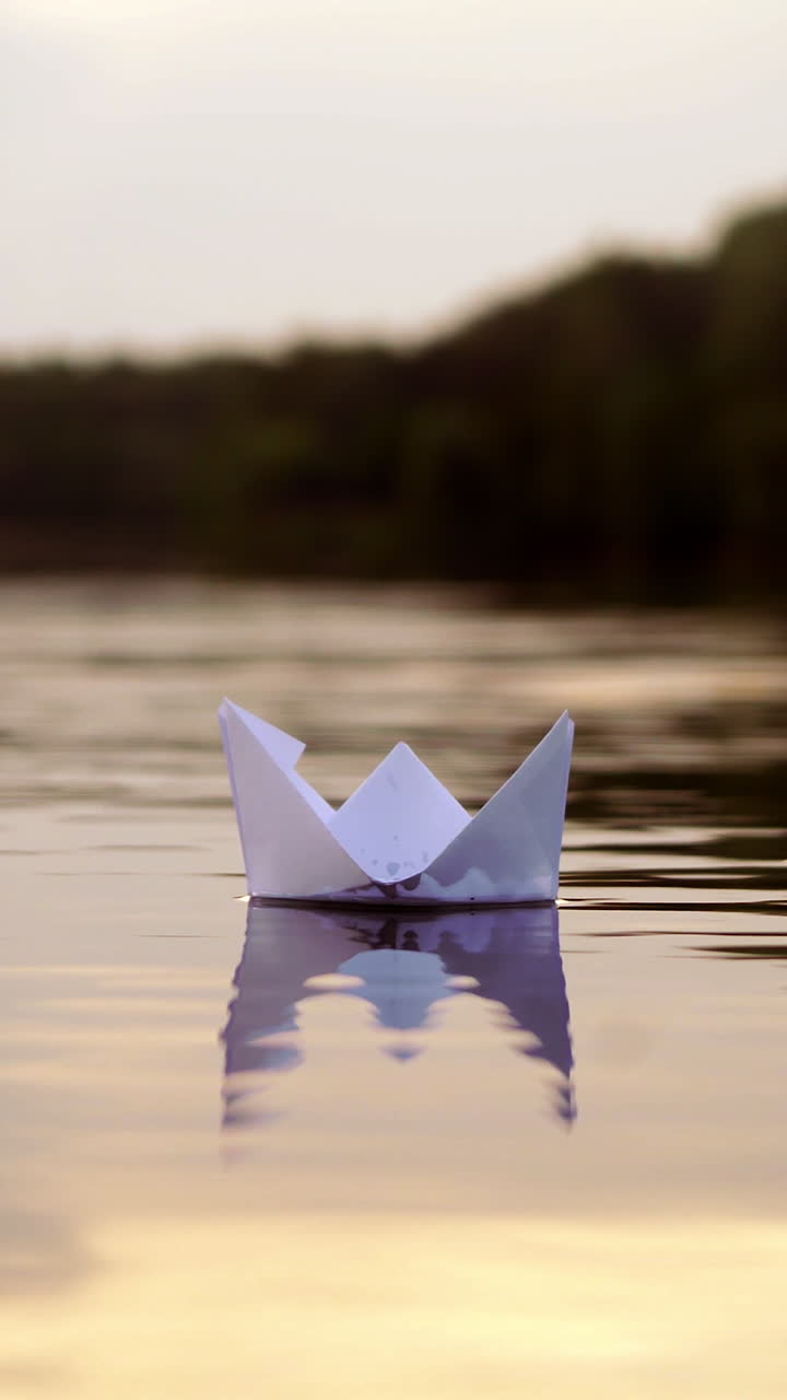 White paper boat is floating on the water near the forest background in the evening. Origami boat floats away into the distance along the river Vertical video