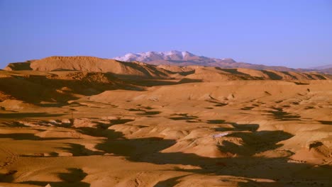 panning medium shot of dry desert landscape with snow covered mountains in the background in morocco