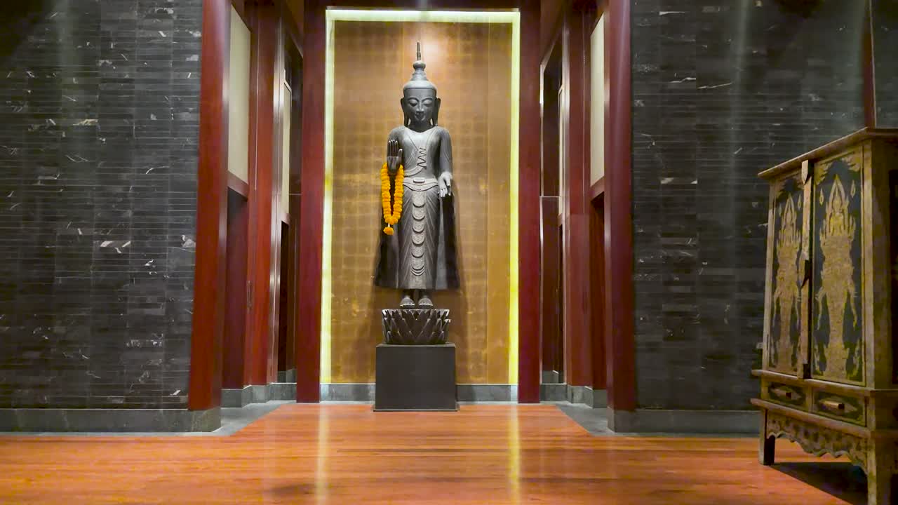 Uno zoom lento verso una statua del Buddha in piedi all'interno di una lussuosa hall d'hotel con pavimento in legno.