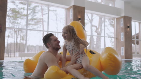 Una-Niña-Alegre-Se-Ríe-En-La-Piscina-Sentada-En-Un-Divertido-Flamenco-Inflable,-Papá-Sostiene-A-Su-Hija