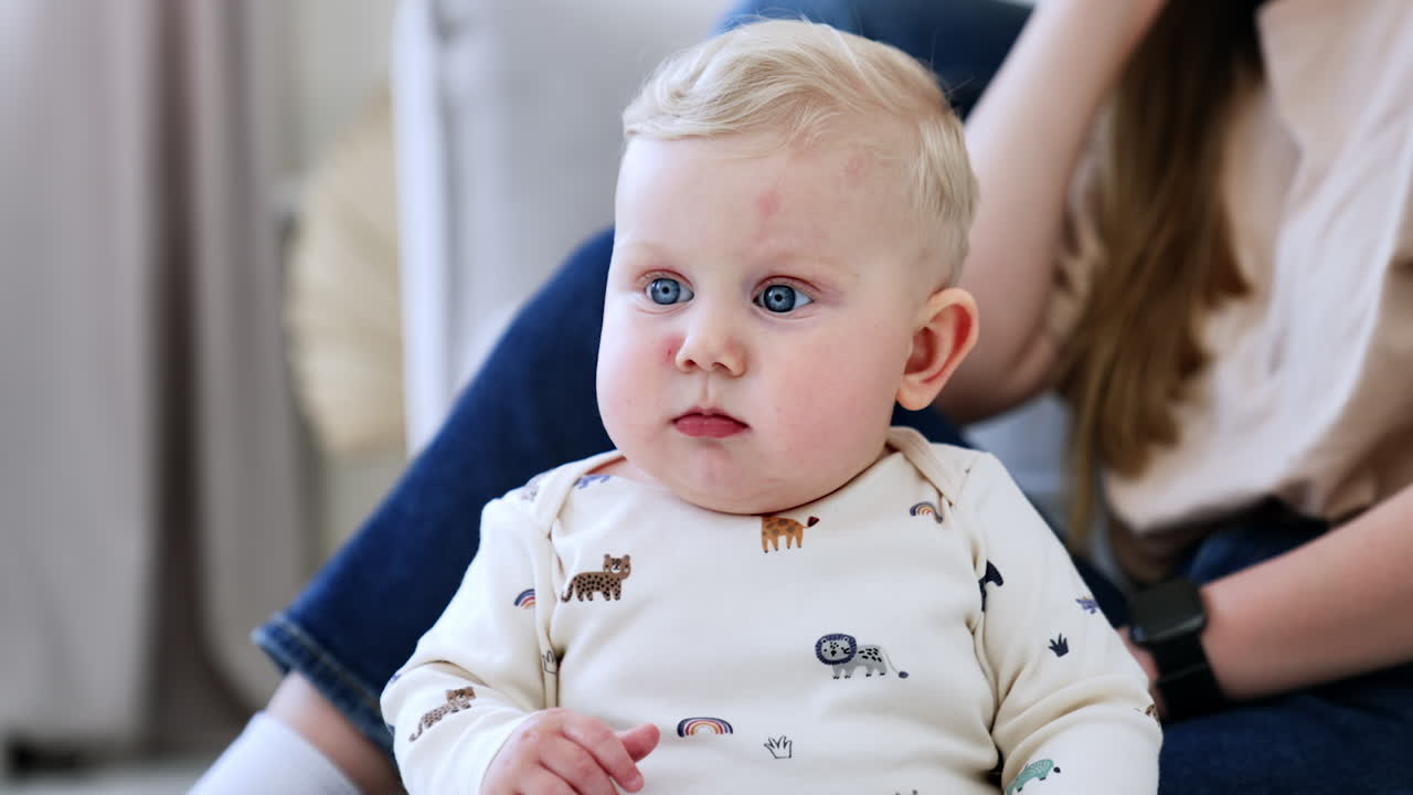 Adorable chubby blond baby is sitting calm. Close up portrait. Unrecognized parents at backdrop.