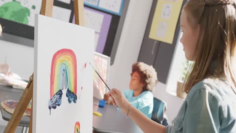 video of happy caucasian schoolgirl standing at easel painting in art class, copy space