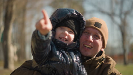 a close-up of a father and son sharing a happy moment outdoors, the child wearing a shiny black jacket, excitedly points at something while smiling widely, the father, also smiling