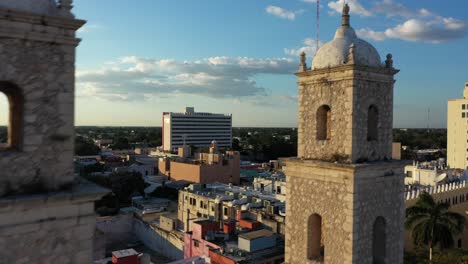 Aerial-camera-at-sunset-slipping-to-the-right-and-rising-behind-the-bell-towers-of-Rectory-Jesus-in-Merida,-Yucatan,-Mexico