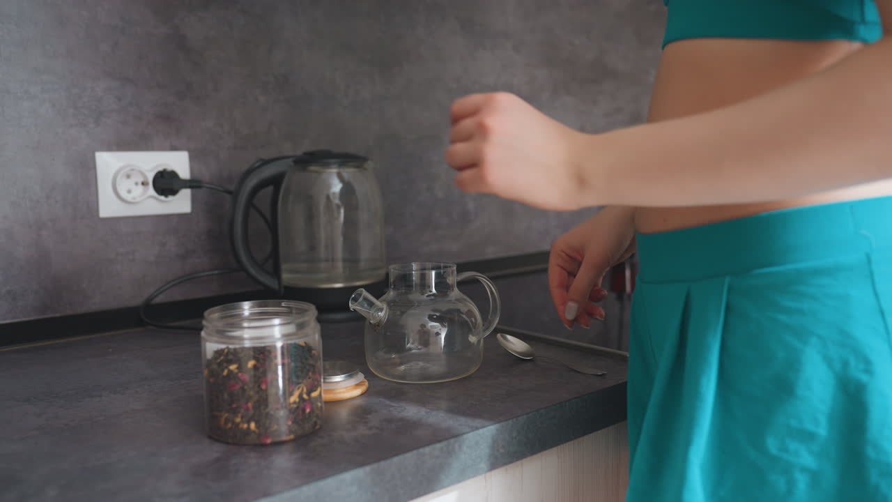Caucasian Woman Scoops Tea Leaves Into Glass Teapot On Modern Countertop With Kettle In Background, Relaxed Morning Ritual, Steam Rising, Aromatic Herbs, Careful Hands, Casual Homewear