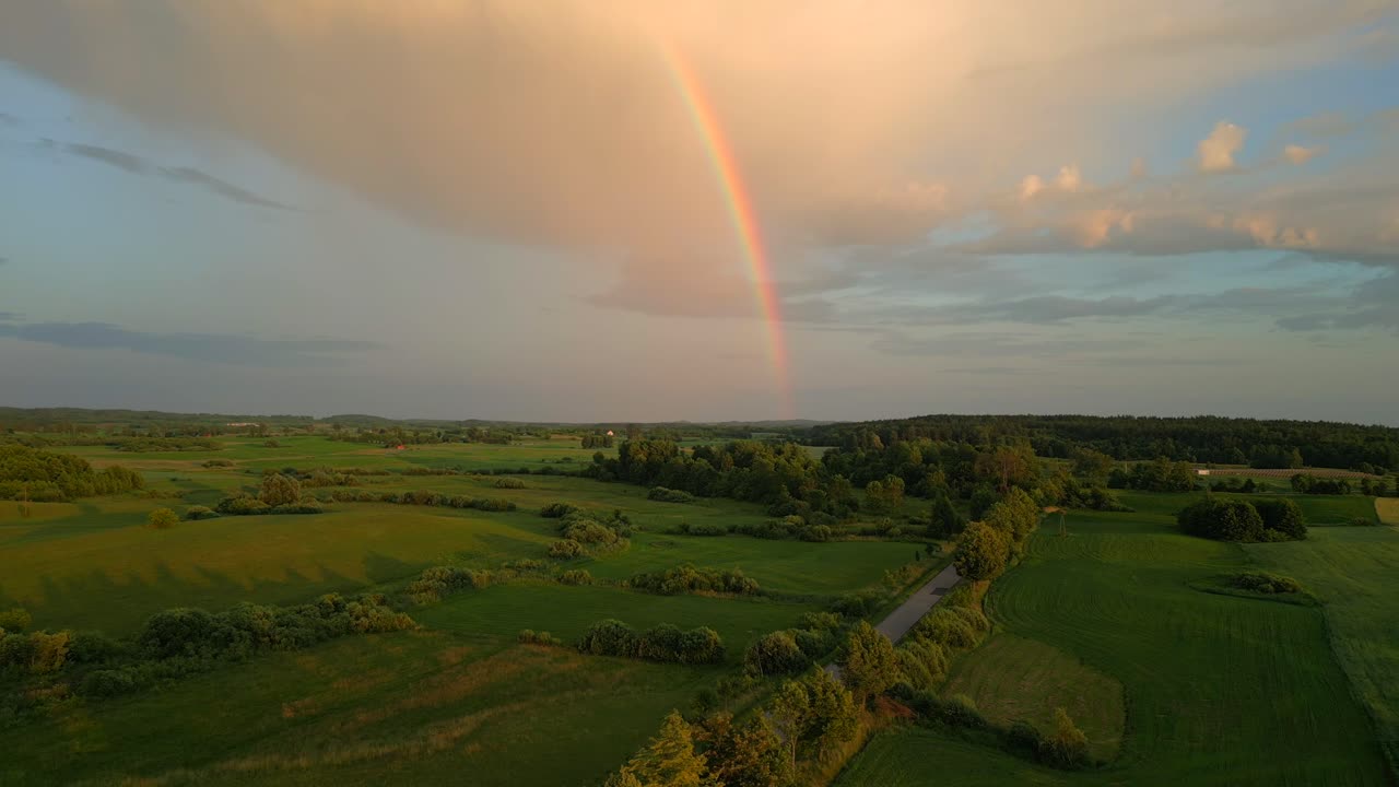 puesta de sol sobre un paisaje con campos y una carretera sobre la cual se levanta un arco iris
