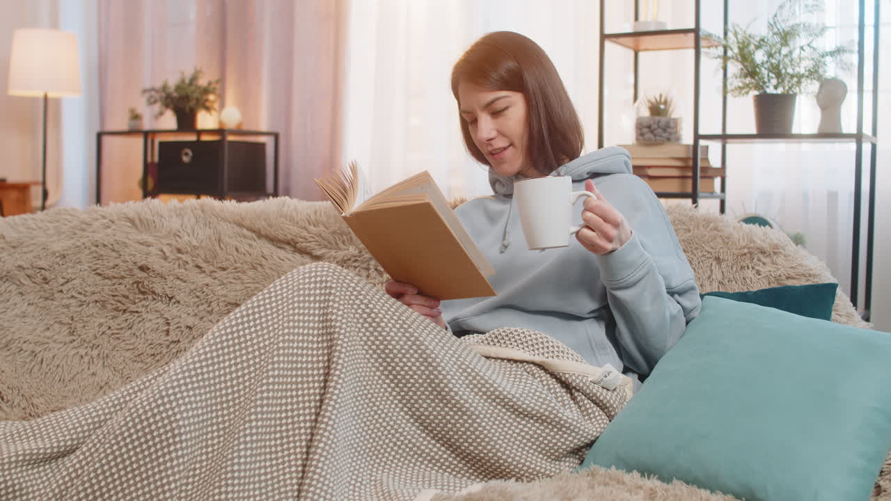 집 의 소파 에 핫 초콜릿 한 잔 을 마시면서 흥미진진 한 책 을 읽는 미소 짓는 젊은 여자