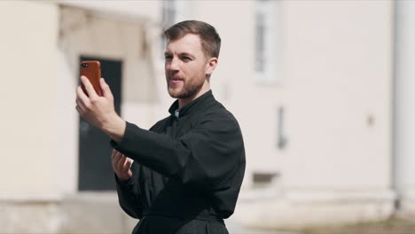 Un-Joven-Sacerdote-Agradable-Está-Hablando-En-Una-Videollamada-A-Través-De-Un-Teléfono-Inteligente