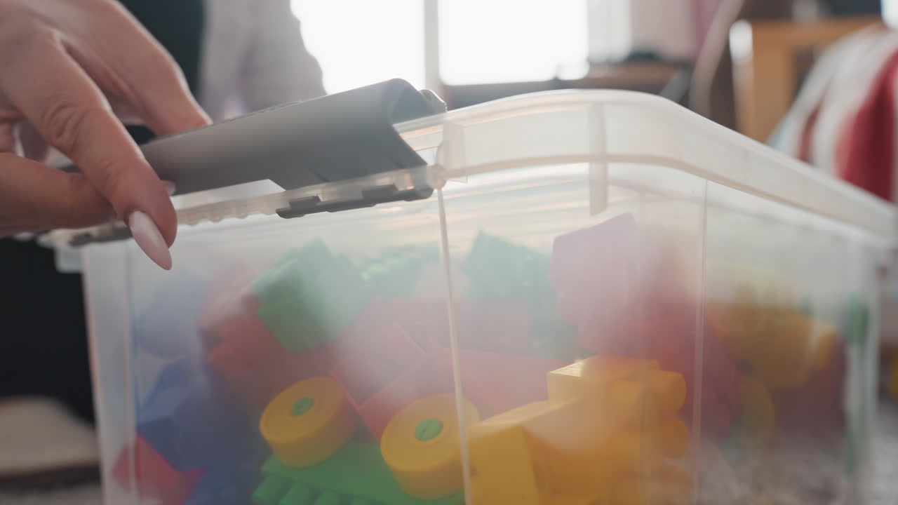 Lowangle toy box on carpet, mother kneeling and unlatching lid while son watches, colorful blocks piled inside translucent container, warm backlight, action of reaching and gripping latch, lively