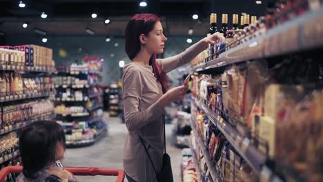 Young-beautiful-female-costumer-is-choosing-spices-in-a-local-supermarket.-Her-toddler-son-waits-for-her-patiently-in-a-push-cart.