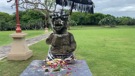 Tilt-up-to-a-stone-God-statue,-flowers-and-offerings-in-front-of-the-figure-in-a-green-lush-park