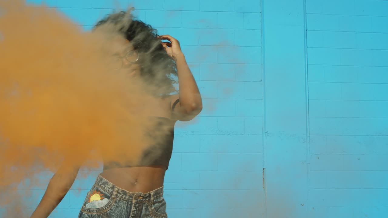 hermosa mujer joven sosteniendo colorida granada de humo bailando afuera contra la pared azul