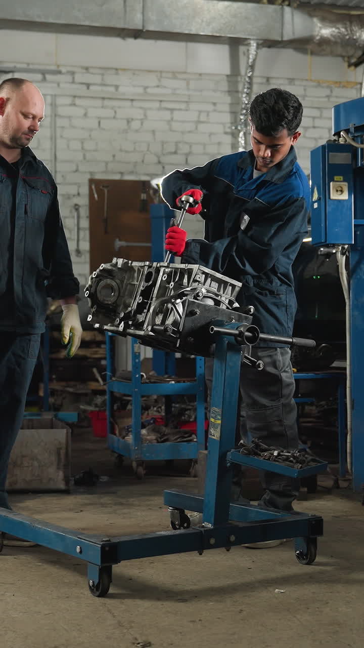 dos ingenieros en un taller automotriz, uno con guantes rojos trabajando en un motor de coche mientras el colega observa, el fondo presenta coches estacionados, herramientas mecánicas y varios equipos