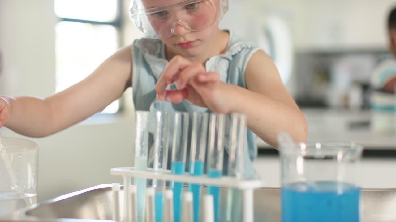 Holding pipette, girl with goggles pipetting blue liquid across tube rack at counter for science