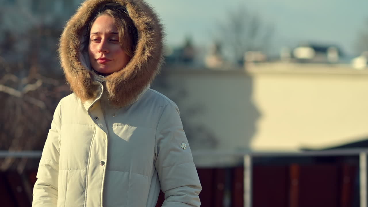 A woman stands outside on a bright winter day, dressed in a warm, fur-lined jacket. She enjoys the sunlight against a backdrop of modern buildings and bare trees