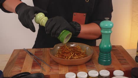 Slow-motion-shot-of-chef-hands-wearing-black-rubber-gloves,-grinding-salts-into-a-plate-of-tasty-crispy-pai-potatoes-from-mill,-to-make-sure-the-dish-is-properly-seasoned-with-salt-and-pepper