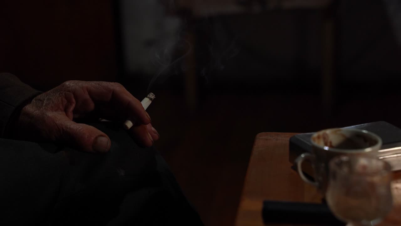 mano de anciano sosteniendo un cigarrillo cerca de una taza de café en una habitación oscura, anciano