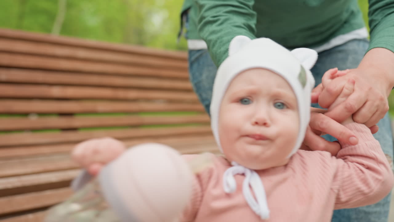 Older Person Aids Baby Standing On Seat, Elderly Individual Assists Toddler To Stand On Park Bench, An Aged Person Supports Young Child As They Stand On Park Bench Surrounded By Greenery