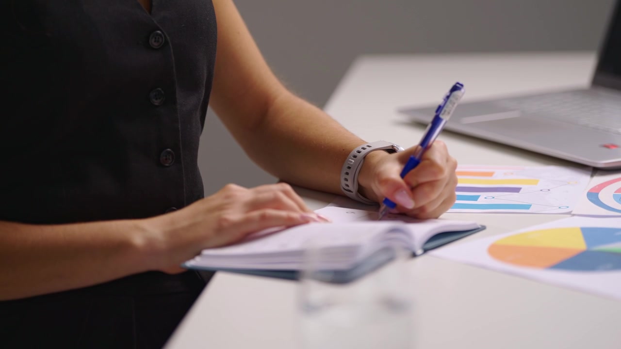 Profilo laterale di una dietologa concentrata che guarda in basso e scrive su un'agenda alla sua scrivania, con laptop e tabelle nutrizionali nelle vicinanze durante una consulenza sanitaria professionale e la pianificazione dei pasti.
