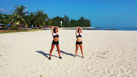 happy healthy girls with beautiful fitness bodies exercising on white sand area of exotic beach near tropical resort with blue sea background in thailand