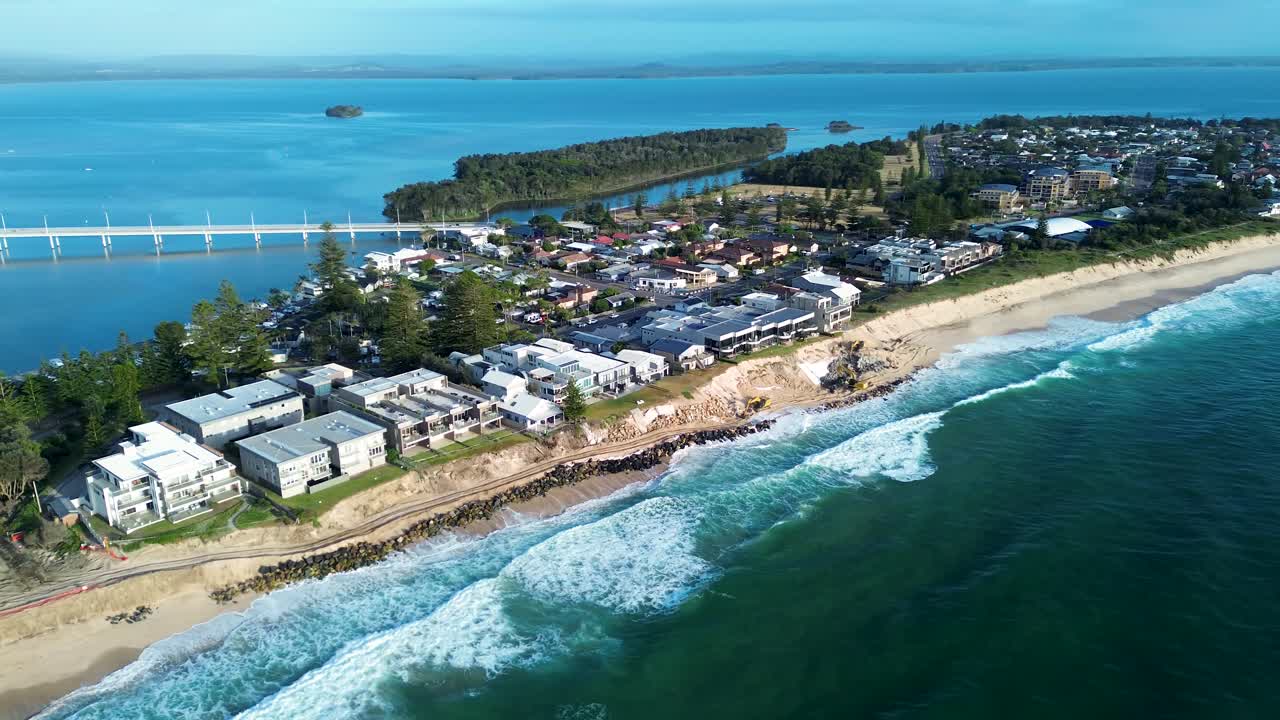 Drone aerial landscape of ocean waves crashing on beach rocky break wall with suburban town housing and The Entrance bridge in the background Tuggerah Lake NSW Central Coast Australia tourism travel